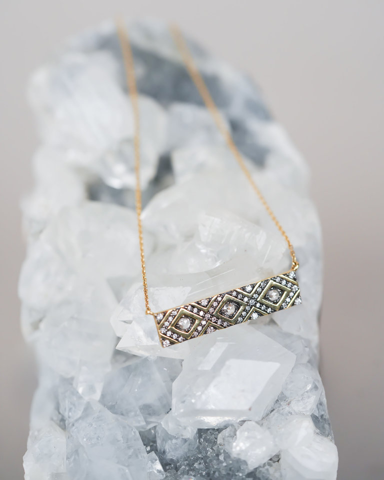 A crystal holds a horizontal bar necklace. The necklace has a zig zag pattern of diamonds referencing the rattlesnake in the name.
