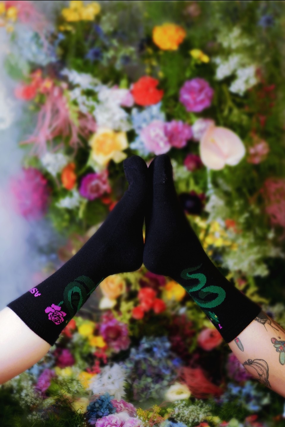 Two feet come together sole to sole showing the Asrai sock in black with pink Asrai Garden text and rose plus a green snake.