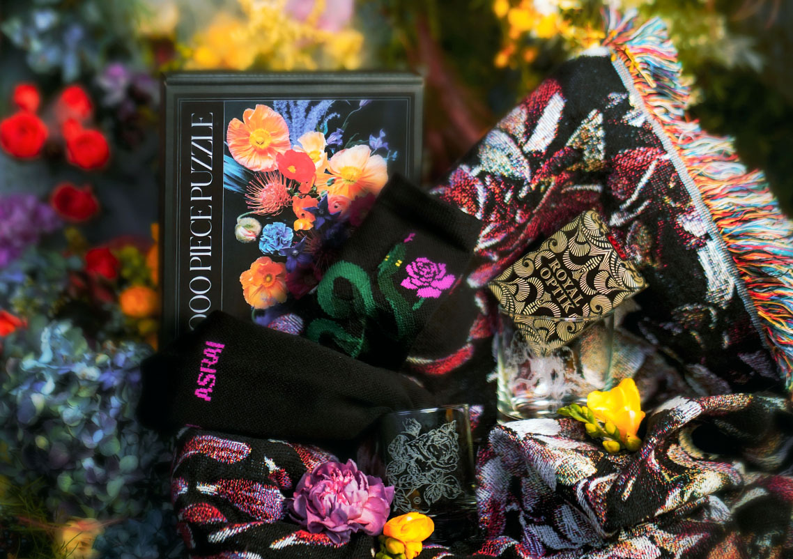 A colorful floral background showcases Asrai Garden''s gift set for the holidays. The gift set features an Asrai puzzle, snake socks, Uusi playing cards, etched numbers and floral blanket
