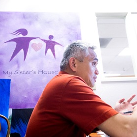 A man sits at a desk, gesturing with his hands in conversation, before a purple banner that reads My Sister''s House