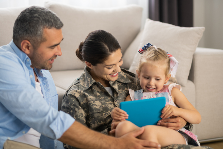 Military family smiles together with child looking at tablet.