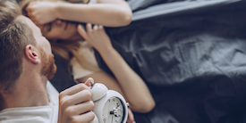 Man holds alarm clock above woman who is in bed - image