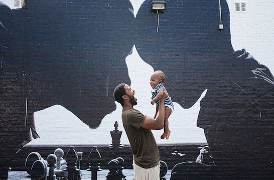 A Black man is holding a baby up in the air in front of a painted mural with a father and son.