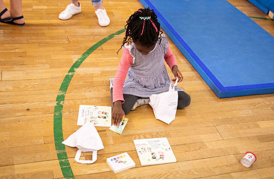 Literacy. Small Black girl with braids wearing a pink top and blue strip dress kneels on gym floor around papers.