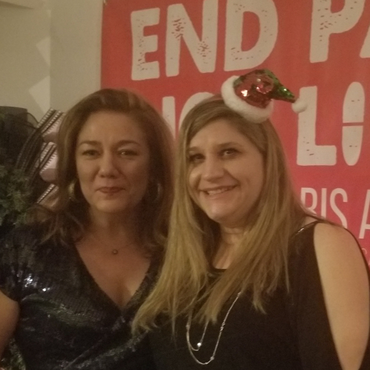 Steph Sherer and Debbie Churgai dressed formally and posing next
to one another. Debbie is wearing a small sequined santa hat. They are
standing in front of a red banner that reads \