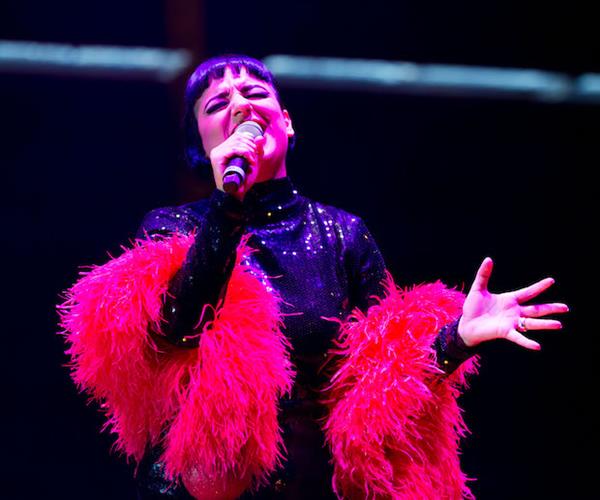 Performer singing in microphone in sequinned black top and red fluffy scarf.