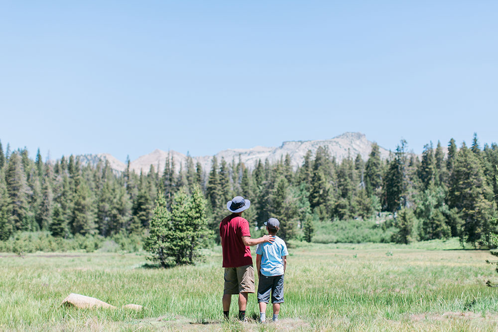 A dad and son in hats look out over a montain meadow
