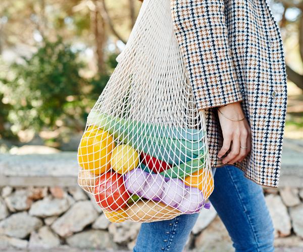 a reusable bag filled with colourful fruit and vegetables is slung over a woman's arm