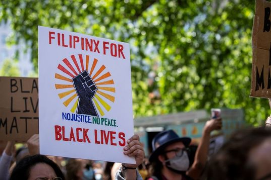 A protester holds a homemade sign that says, "Filipinx For Black Lives No Justice No Peace" with a black power fist and rainbow coming out from the center of the fist 