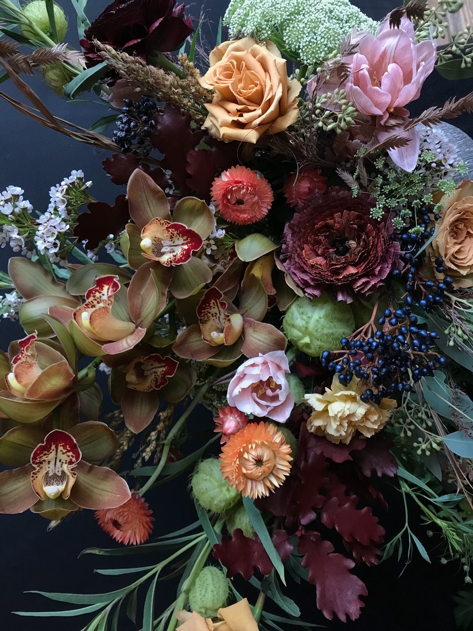 An explosion of dark and neutral florals fill the photos space. Tan Cymbidium orchids mix amongst peach Strawflower, white Queen Anne''s Lace, burgundy Ranuculus, blue berries and Milkweed pods.