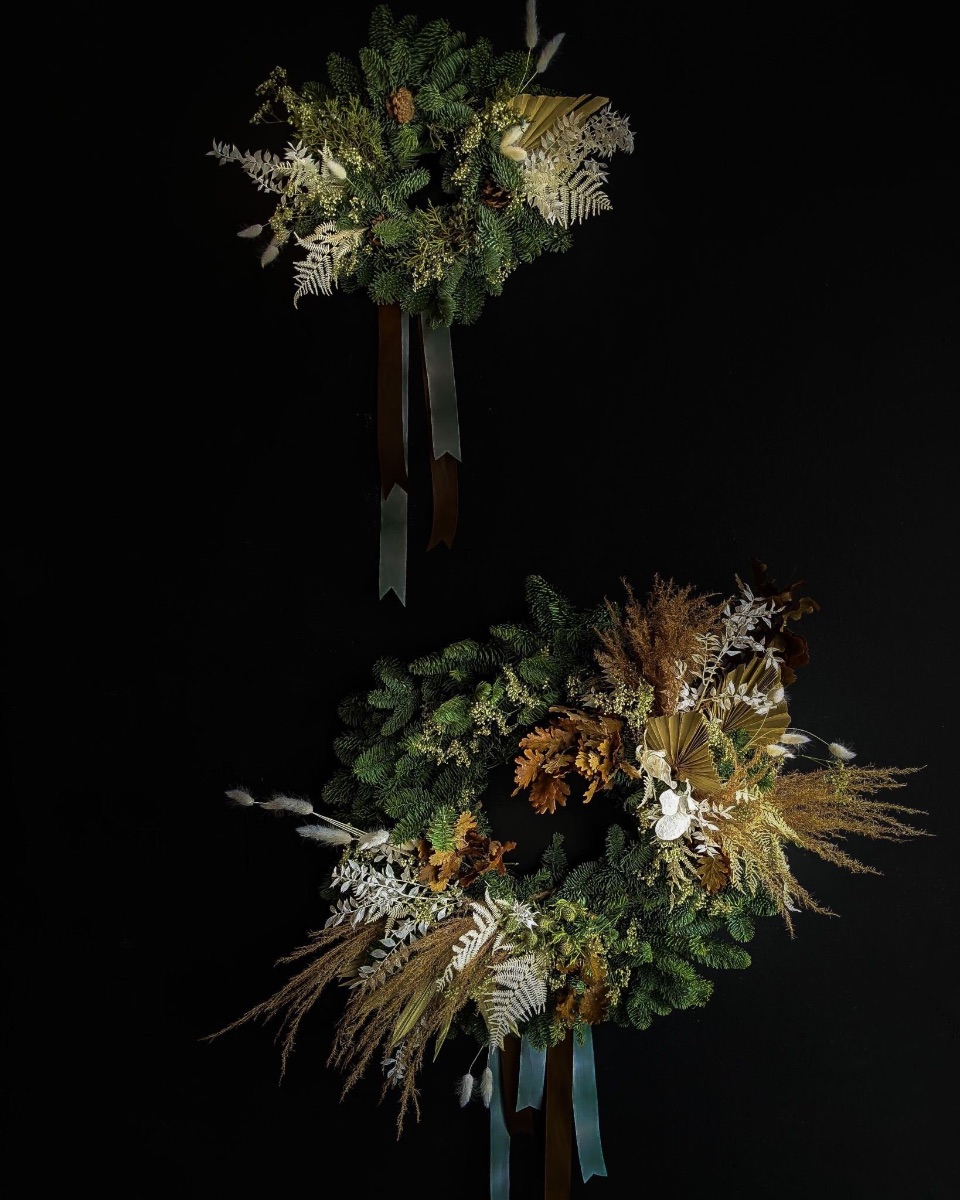 A small wreath hangs on a black wall over a large wreath. The wreaths'' base are traditional holiday greens but amplified Asrai style with dried palm fans, grasses and bleached fern.