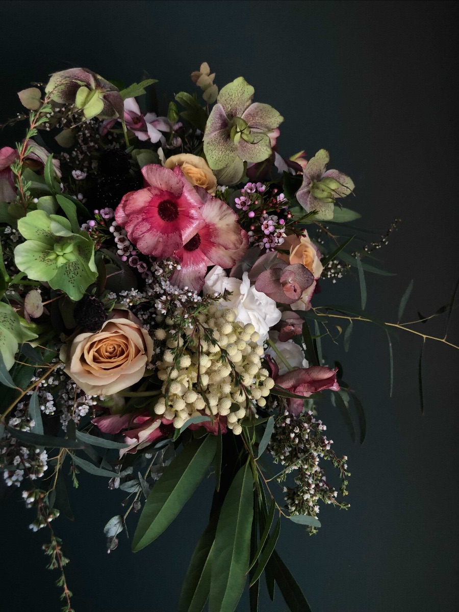 A bouquet cascades out of the left side of the photo. Green and purple Hellebores mix with golden roses and burnt red Butterfly Ranunculus.