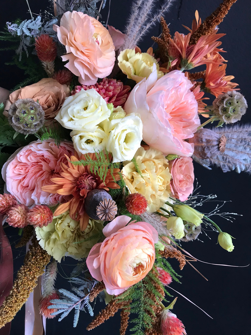 A summery peach explosion of a bouquet. Peach Garden Roses and Ranunculus take center stage surrounded by poppy pods, spray Mums and fuzzy grasses.