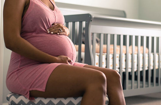 Black mother. Pregnant Black woman wearing pink dress holding her belly in a room with a white crib.
