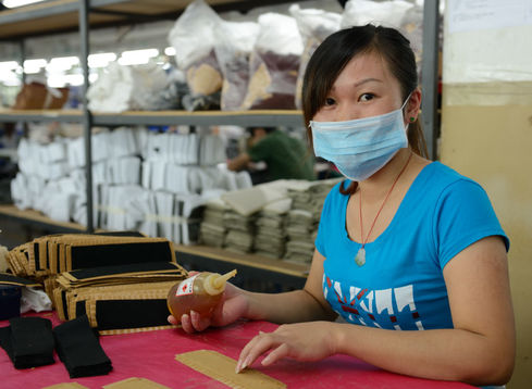 Young Chinese woman seated in a factory with a blue t-shirt, blue face mask, glue in one hand, and fabric on a table.