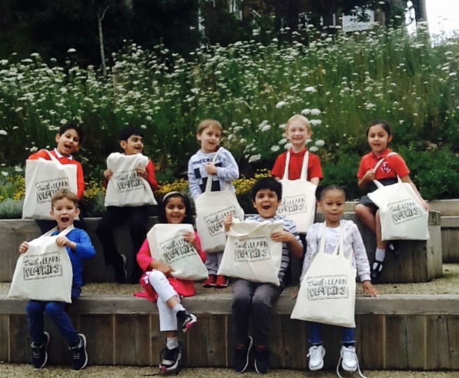 A group of smiling children sit on some steps and hold up cream tote bags