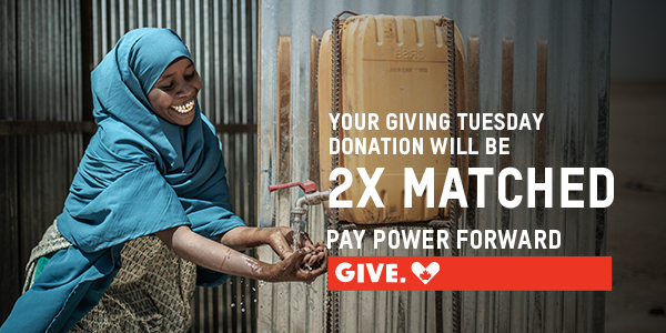A woman with a blue head scarf is smiling and washing her hands from a jug. Text reads: Your giving Tuesday donation will be 2x matched. Pay Power Forward. Give.