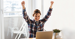 happy woman raises her arms in celebration as she looks at her computer screen