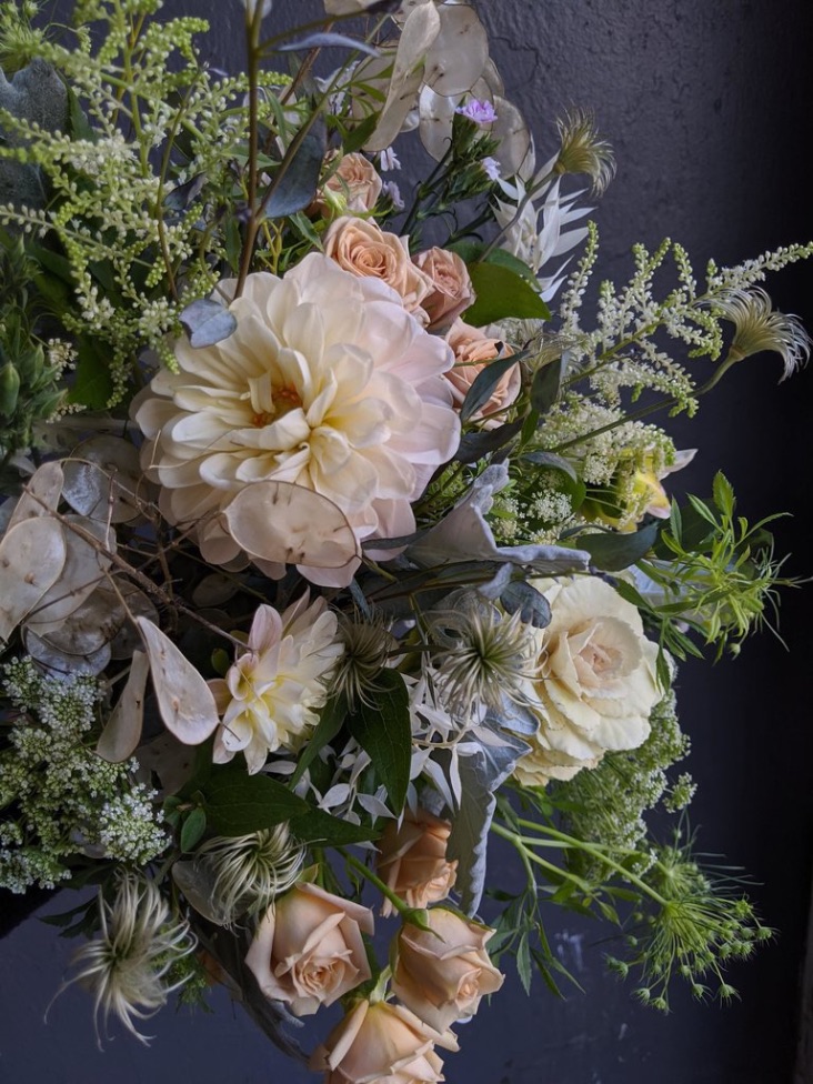 Peachy and tan tones are featured in this Asrai floral arrangement. A large cream Dahlia sits center stage surrounded by white queen anne''s lace, pale cabbage, tan spray roses and preserved lunaria