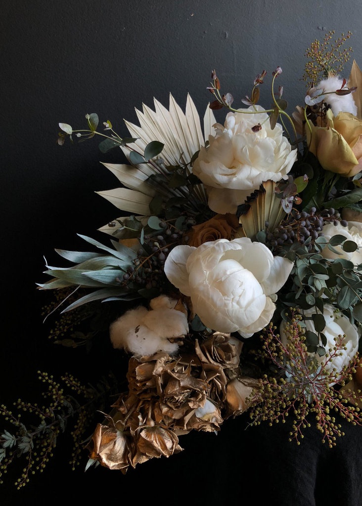 Whites and golds stand as the focal color palette of this Asrai arrangement. Gold painted spray roses cascade out of the side of white cotton and large white peonies. Golden roses and blue berries mix in while a dried palm fan frames the back of the arrangement. 