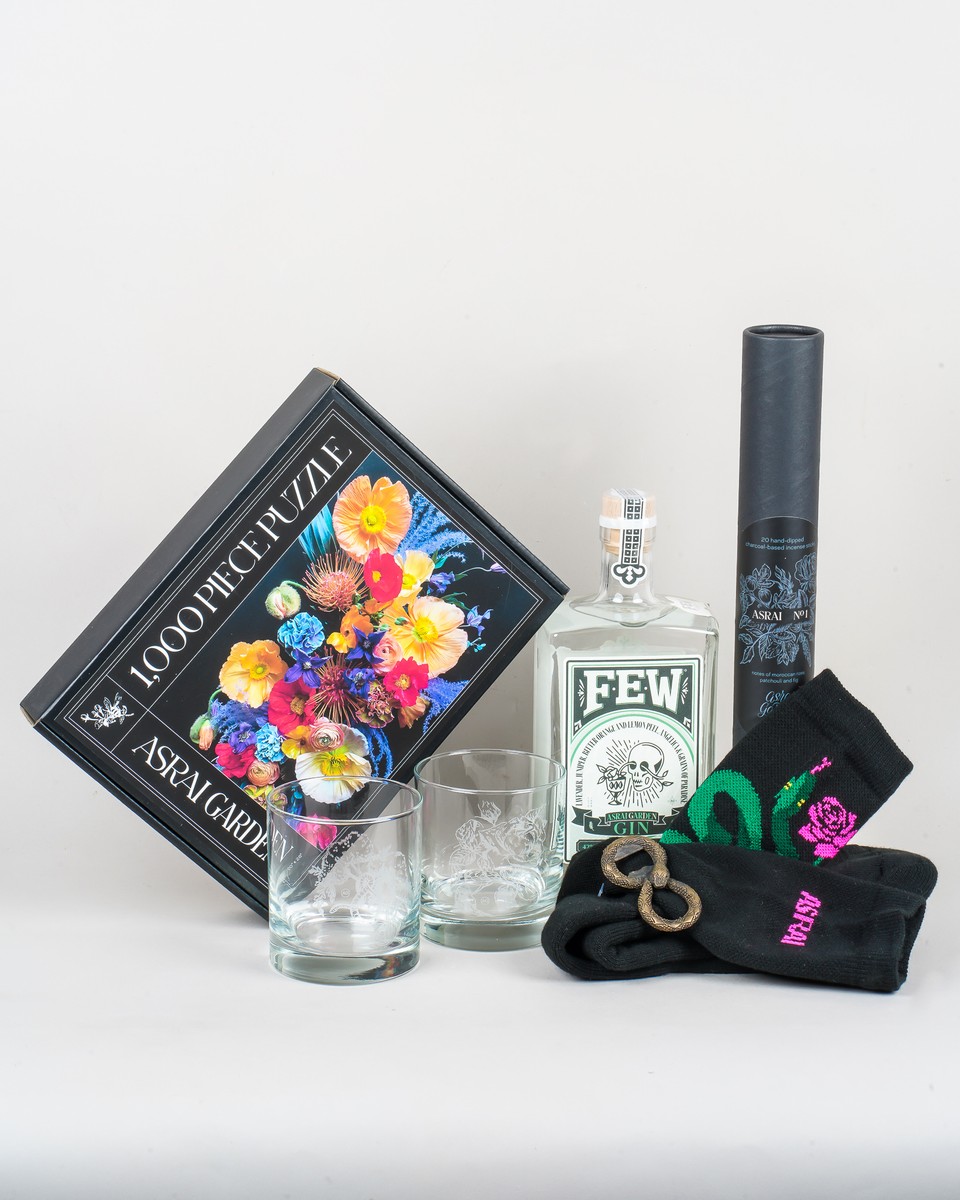 A collection of Asrai inspired gifts featuring a brightly colored floral puzzle, a bottle of Asrai X Few Gin, black Asrai tube socks with snake motif, brass snake bottle opener, two etched rocks glasses and long black tube of incense