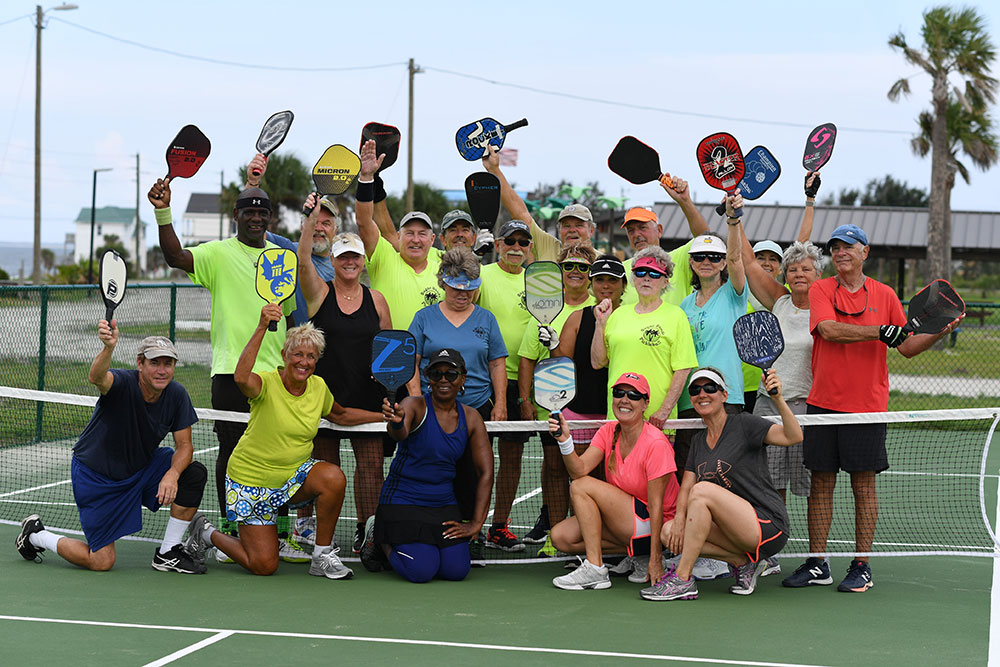 30 people pose for a camera on a pickleball court, holding their paddles aloft