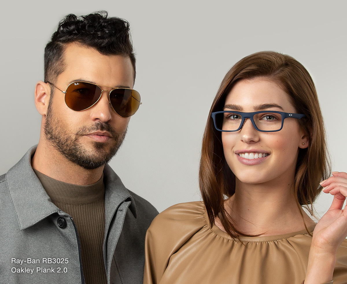 A man with wavy, brown hair wearing a gray jacket over a brown shirt and Ray-Ban RB3025 sunglasses standing next to a brunette woman wearing a tan top and Oakley Plank 2.0 glasses