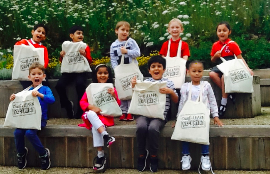 A group of smiling children sit on some steps and hold up cream tote bags