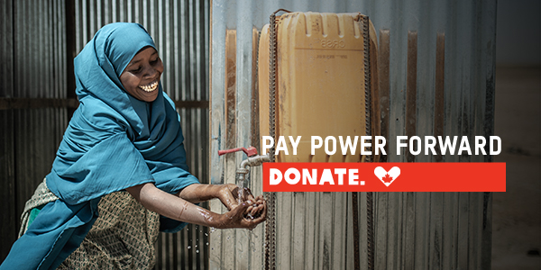 A woman with a blue head scarf is smiling and washing her hands from a jug. Text reads: Pay power forward. Donate.