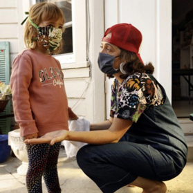 A woman wearing a mask kneels before a young girl, also wearing a mask