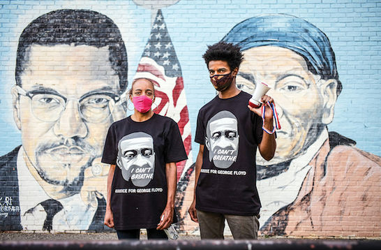 Two protesters stand in front of a painted mural of Malcolm X and Harriet Tubman.