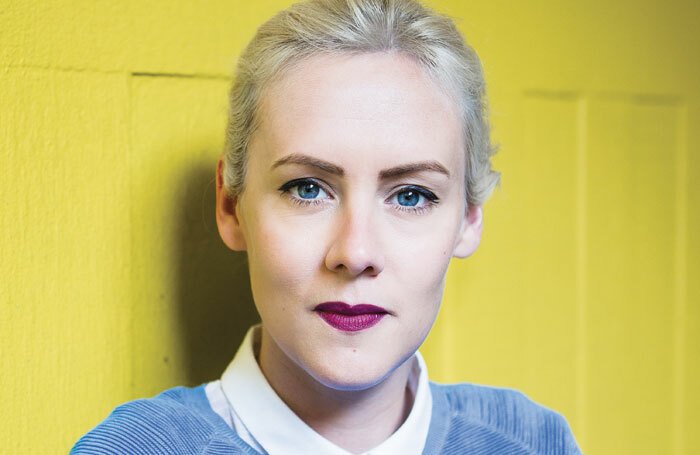 A woman with platinum blone hair in a blue jumper looks directly into the camera, against a yellow background