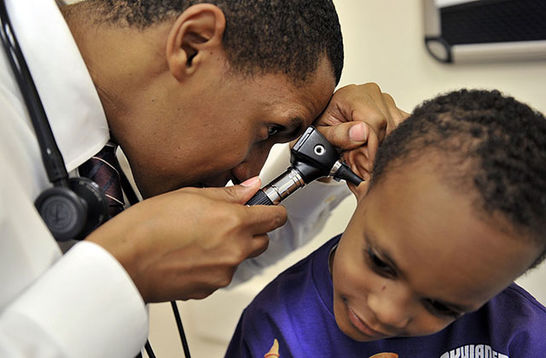 Pediatrics. Black male doctor wearing white coat looks into the ears of a Black male child.