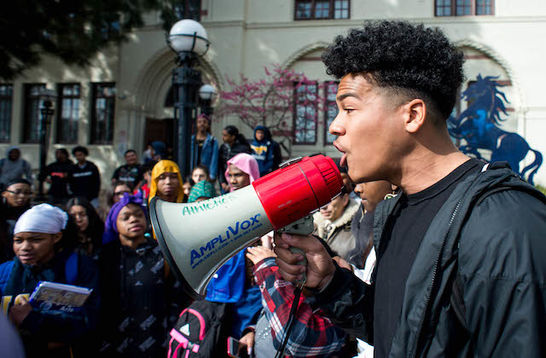 A Black student is holding a megaphone leading a crowd of students in protest.