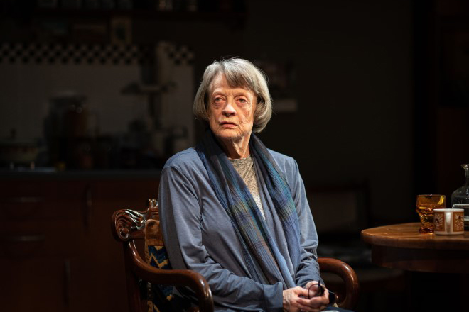 A grey-haired woman looks wistfully out of shot. She sits in a chair, holding a pair of glasses in her hand. There is a table in the right side of shot.
