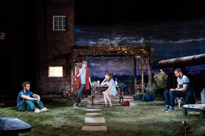 A garden. in the background, a house, a gazebo strung with fairly lights. A large metal pipe runs along the right side of the shot. In centre shot, a woman stands, talking to a young woman sitting at a picnic bench. Two young men sit either side of the image - one on the grass, one on a chair.