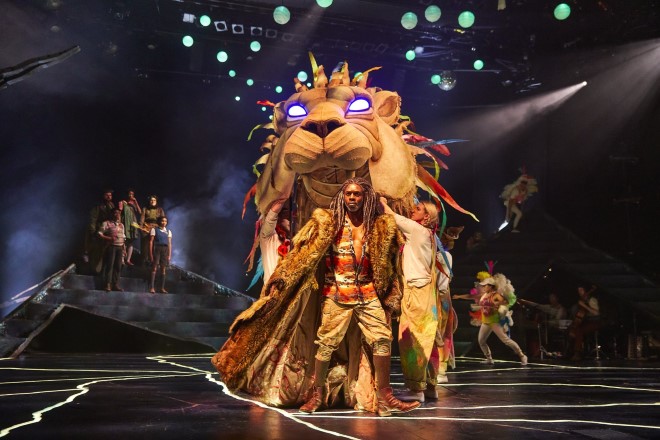 A large puppet lion, led by a man with dreadlocks and flowing robes. In the background, a group of 5 people, out of focus, look on.