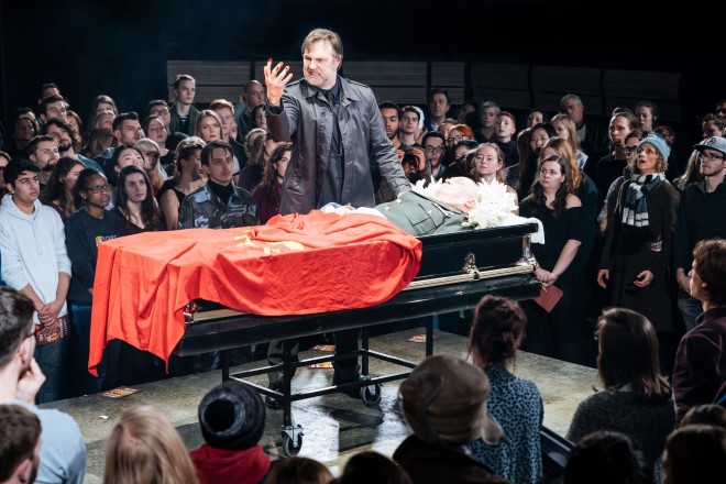 A man in a leather jacket stands over an old man lying in a coffin. He looks angrily at his right hand. The coffin is draped with a red flag. He is surrounded by a crowd, looking on.