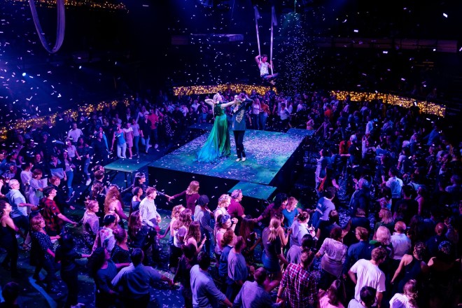 In the centre, a man and woman dance, while a young man suspended in silks looks on. They are surrounded by crowds of audience members. Confetti falls.