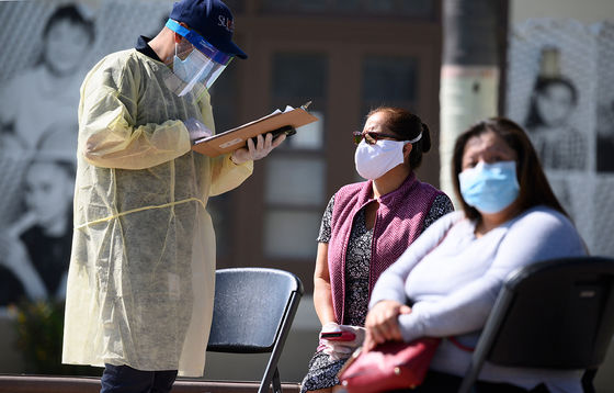 Testing for Coronavirus. A healthcare worker in protective gear, stands and holds a clipboard as two Latinx women sit with masks on  