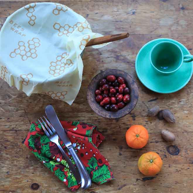 bowl covered in bee''s wrap reusable food wrap on wood table next to cherries, orange, nuts, coffee cup and cutlery