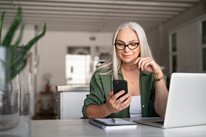 woman looking at phone with computer and notepad