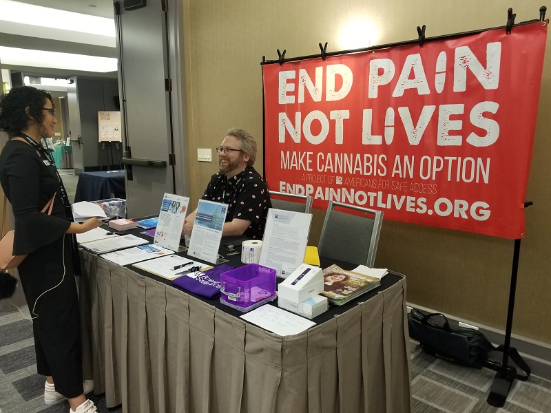 The A.S.A. tabling setup includes a table covered in printed
materials a purple t-shirt and a signup sheet with pens. The man
sitting at the table is in conversation with a woman standing in front
of him. They are both smiling. Behind the table is a large red banner
with white letters reading: "End Pain - Not Lives, Make Cannabis
an Option." The I's in pain and lives are shaped like pills.