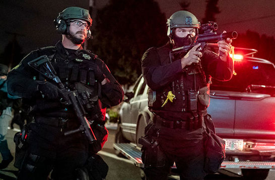 Police Violence. Two officers in uniform wearing helmets and carrying weapons while one is aiming the weapon.