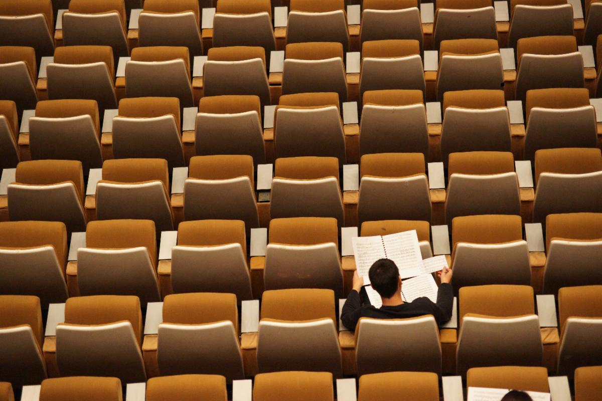Overhead view of empty_ orange seats in an auditorium style classroom. One student in the lower right sits and reads from two open notebooks.