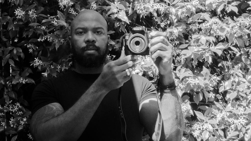 In this black and white photo_ African-American filmmaker Daryl P Jones stands in front of flowering jasmine_ looking directly at the camera. He holds up a digital video camera near his shoulder. He_s bald and has a thick beard.