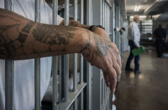 a man with tattoos hangs his arms through the bars of a prison cell. 
