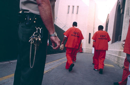 a group of men wearing orange jumpsuits walk away from the camera as a security officer can be seen in the foreground.