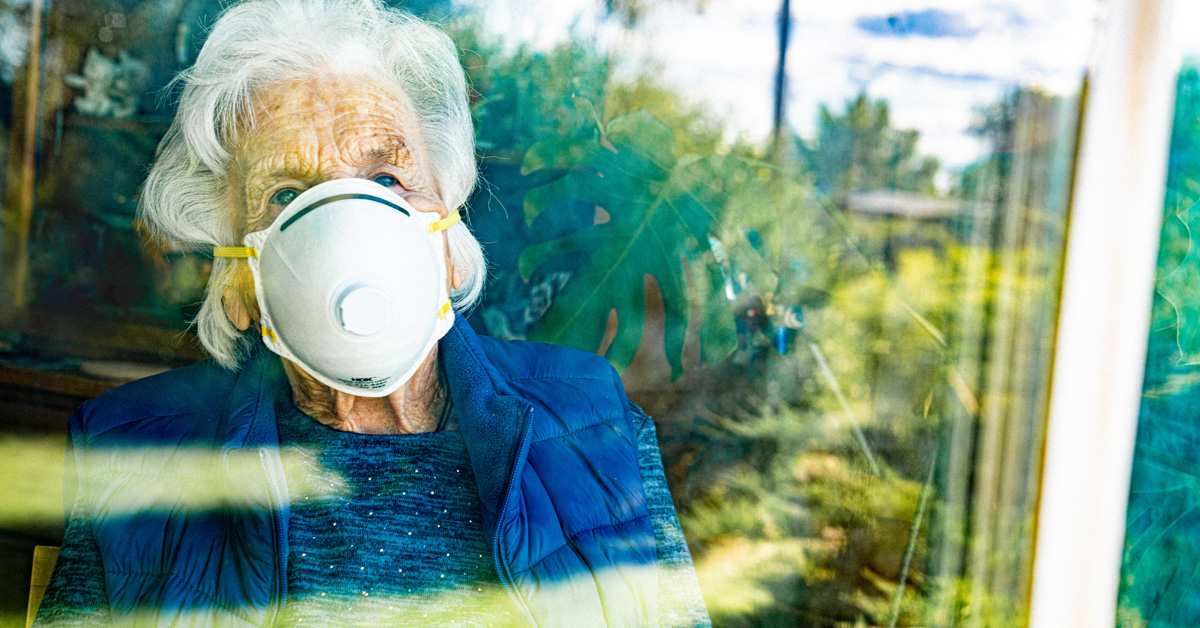 An isolated frail elderly woman in a face mask looks out a window.